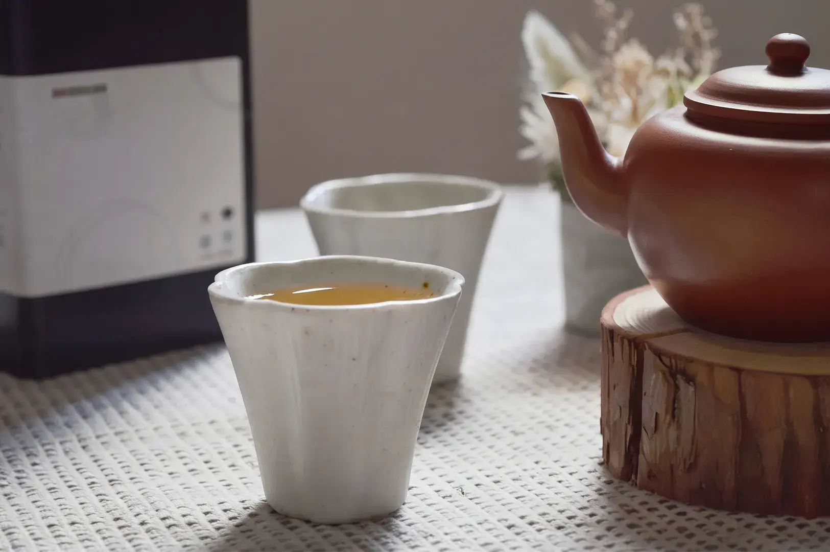 Ceramic teacups filled with tea next to a red clay teapot on a wood slice pedestal.