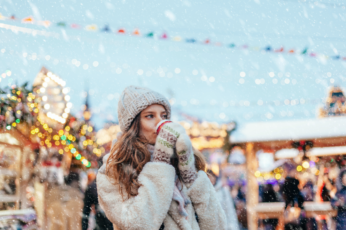 Woman in a winter coat, knit hat, and mittens sipping a hot drink at an outdoor holiday market in the snow during the off-season at the Jersey Shore.