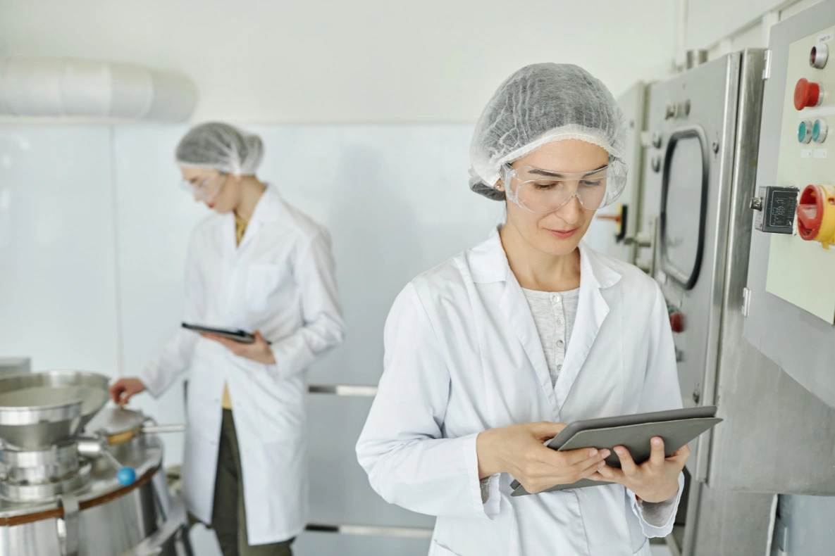 Two people in lab coats and hairnets, one holding a tablet in front of industrial machinery.