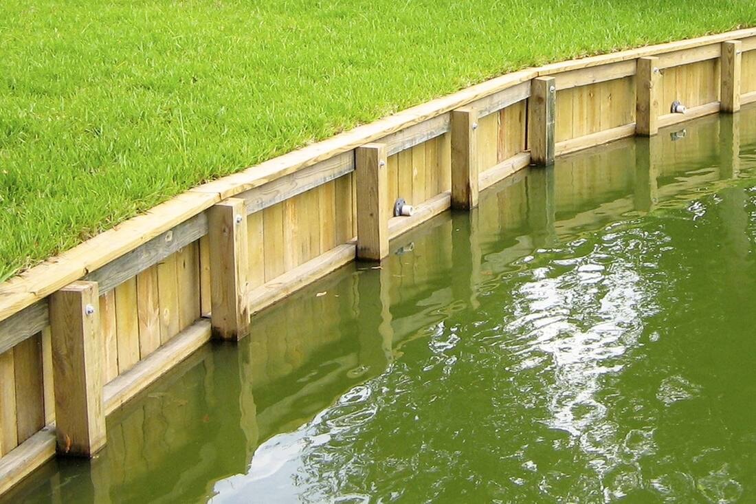 A wooden retaining wall with reinforced posts curves along the edge of a green grassy bank beside calm water.