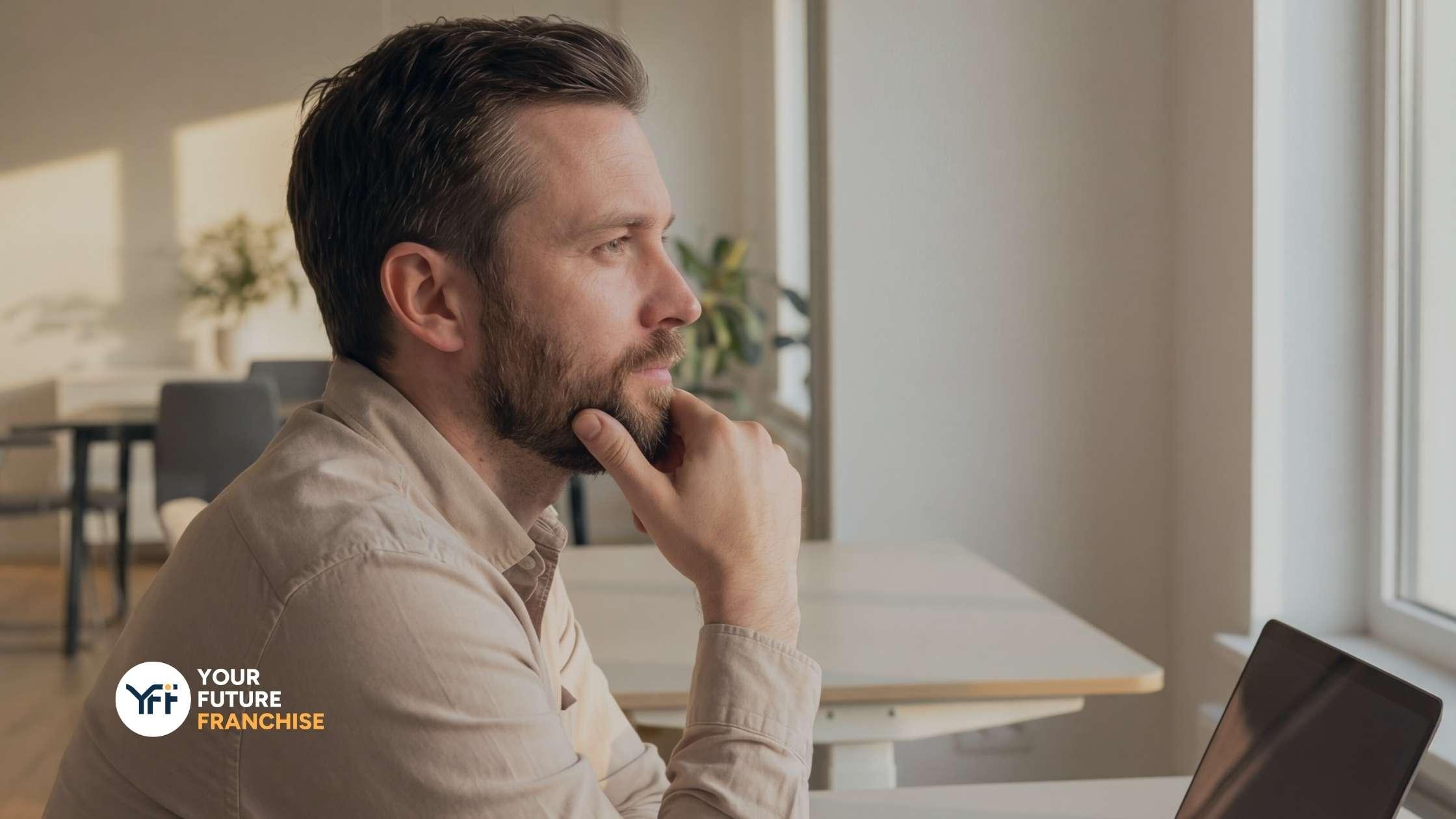 A man thinking and staring out the window with his laptop infront of him