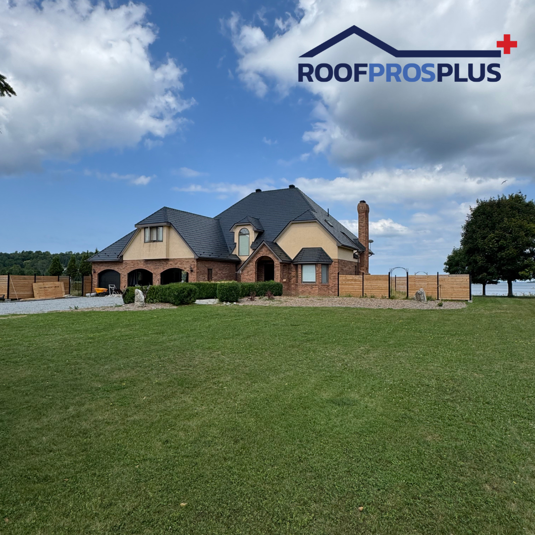 A spacious brick house with dark metal roofing sits on a large lawn under a partly cloudy sky. The RoofProsPlus logo in top right corner. 