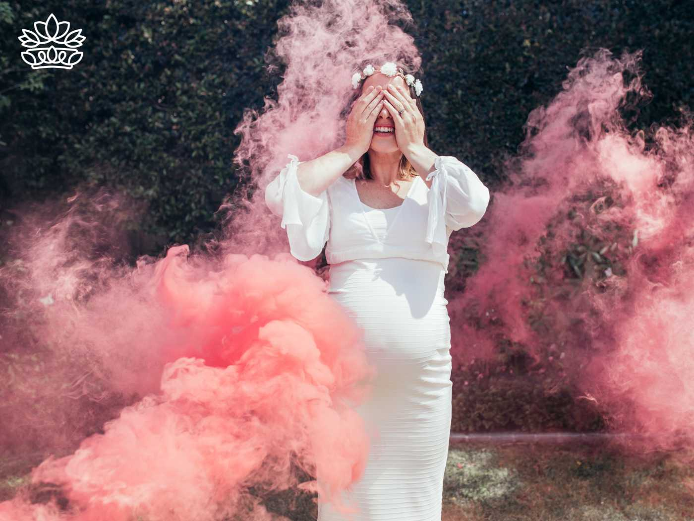 A pregnant woman in a white dress and floral headband stands amidst vibrant pink smoke bombs, expressing joy with her hands covering her eyes, from the Gender Reveal Collection at Fabulous Flowers and Gifts.