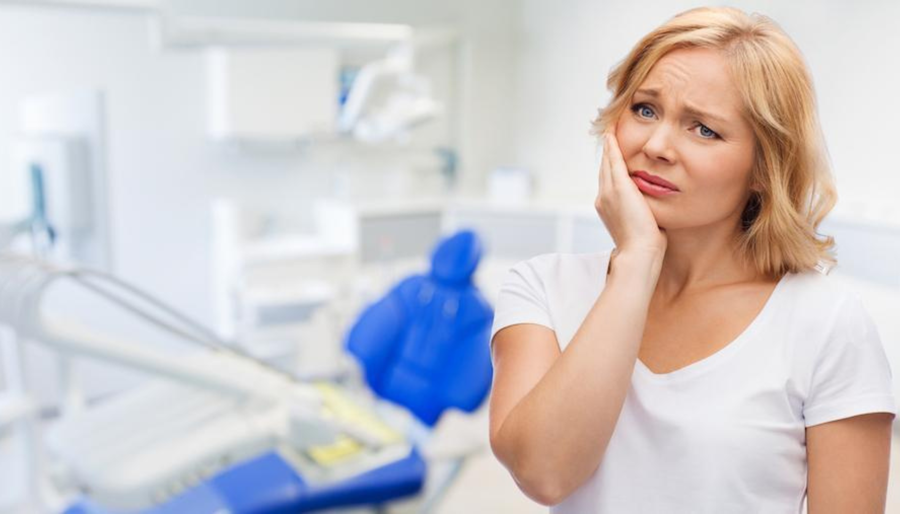 Kvinne i tannlegekontor holder hånden mot kinnet med smertefullt uttrykk, muligens på grunn av tannverk eller kjeveproblemer. 