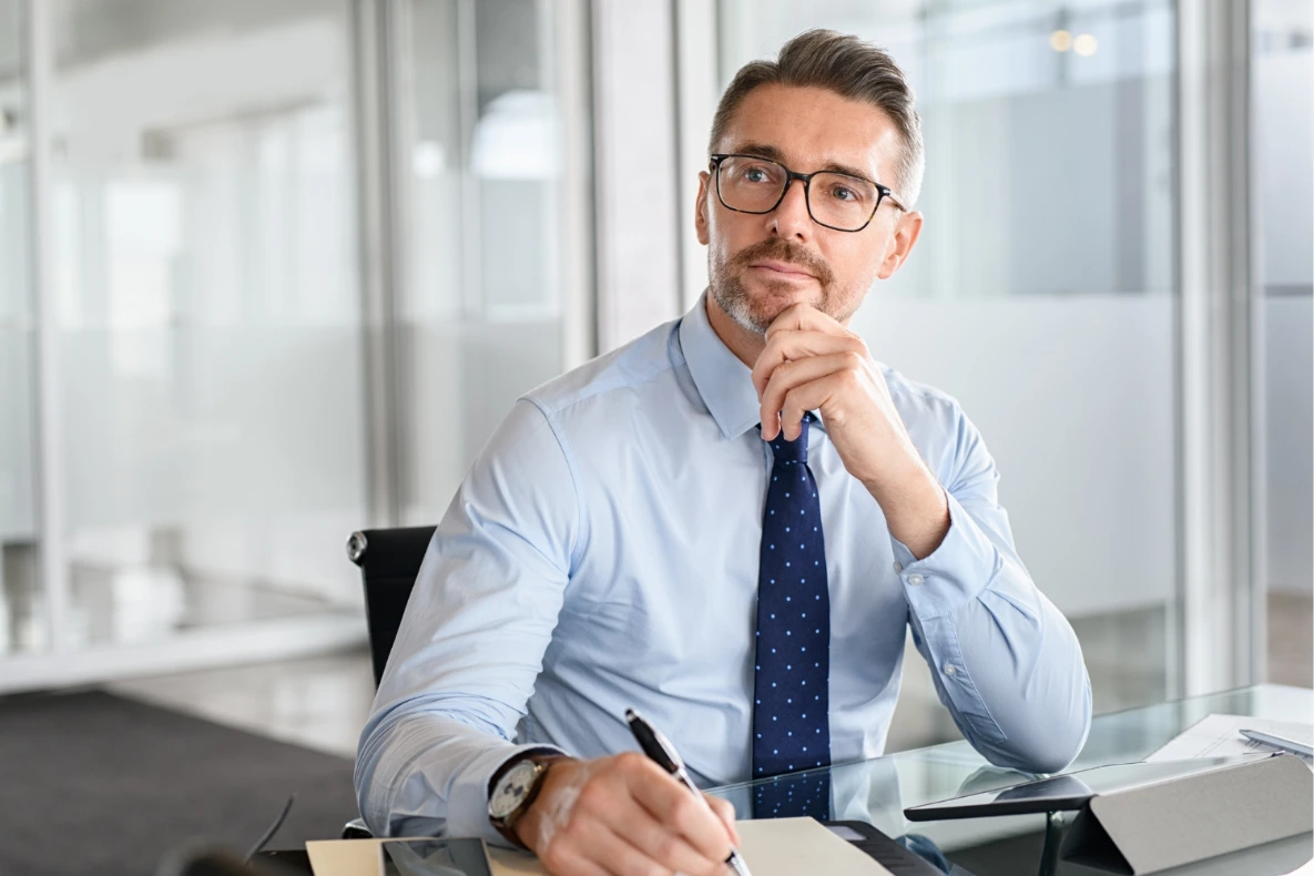A man wearing glasses and a blue shirt with a tie is sitting at a desk, holding a pen and looking away thoughtfully. Shows how a person looks if he wonders about, "How much can I sell my business for?"