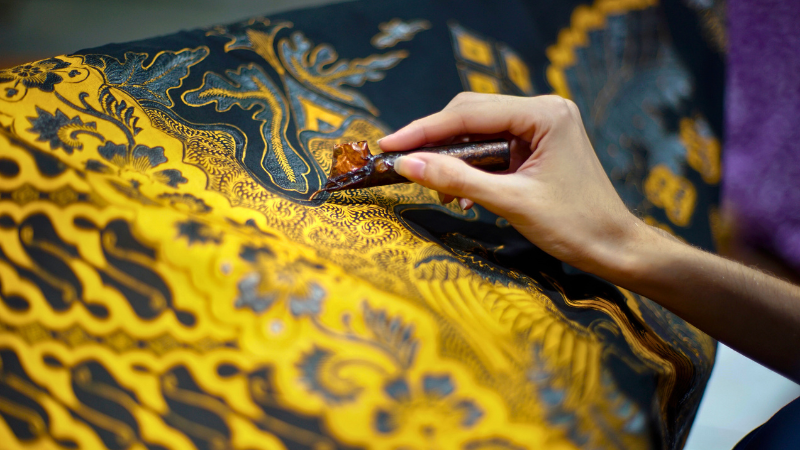 A person is using a pen to write on a piece of Batik Tulis fabric.