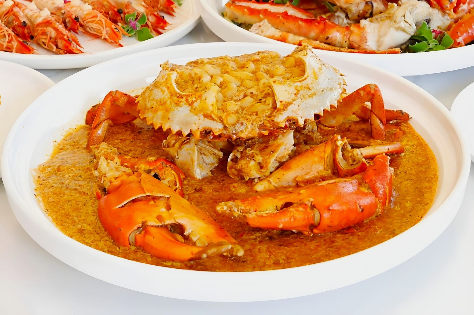 Plate of chili crab with vibrant red claws in rich, spicy sauce. Surrounding plates hold prawns and seafood, creating a festive dining scene.