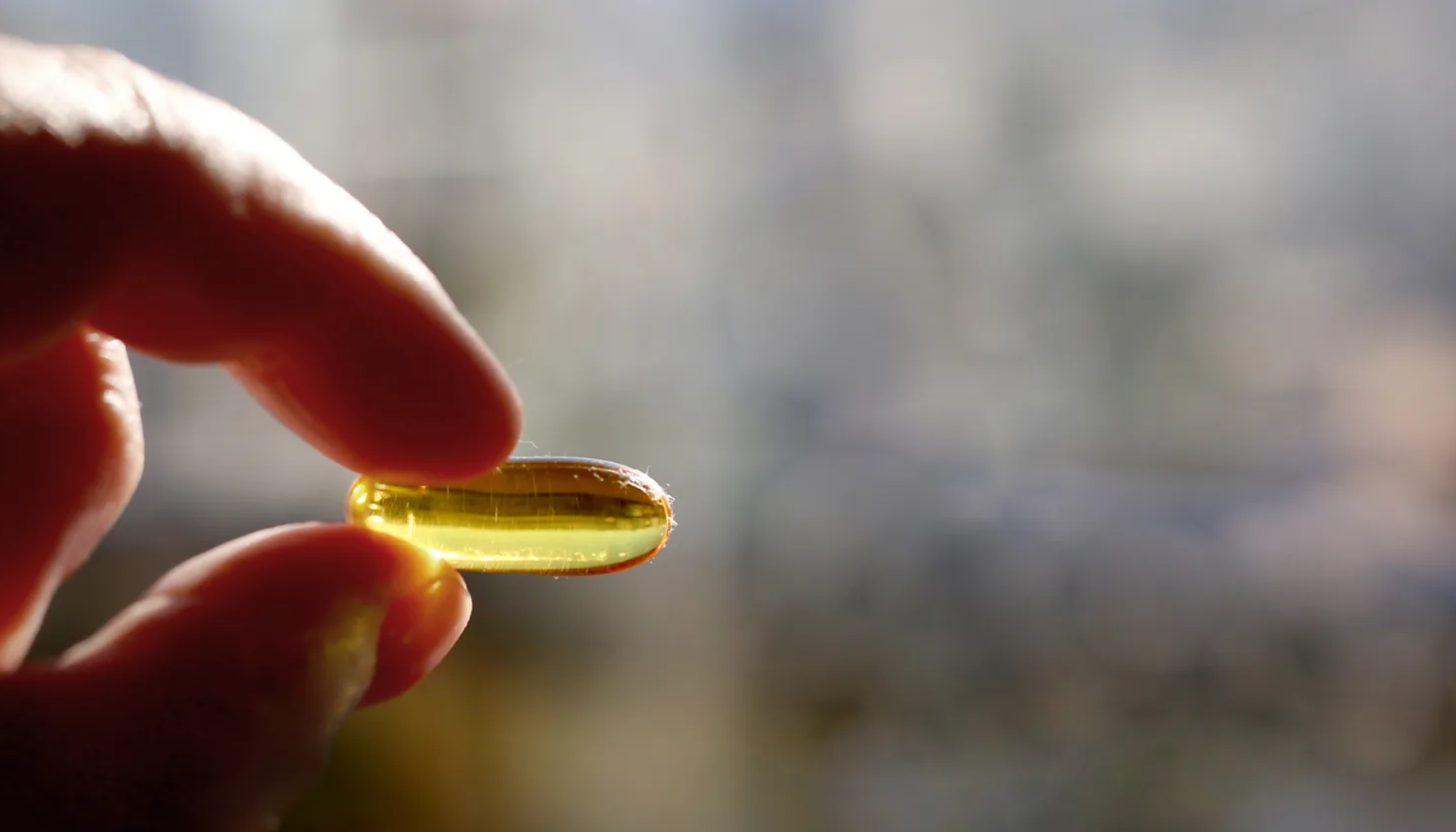 Vitamin D verdier – Hva er normalt og når bør du ta tilskudd? 16 Hånd som holder en gul, gjennomsiktig kapsel mellom fingrene mot uskarp bakgrunn i dagslys.