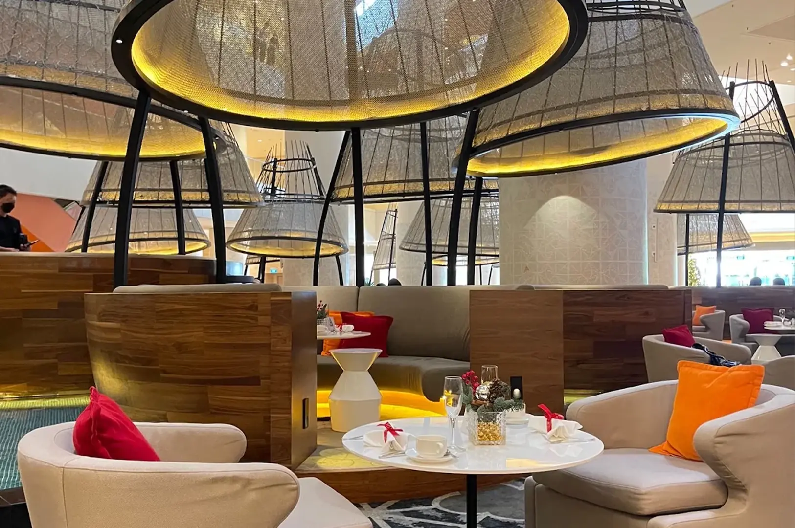 The luxurious atrium of the Pan Pacific hotel features a series of large, bell-shaped, mesh-covered seating pods illuminated by warm interior lighting, creating semi-private lounge spaces over a reflective water feature. In the foreground, a round table is set for high tea with white plates, a teacup, and a glass, flanked by comfortable cream-colored armchairs with vibrant orange and red throw pillows that add a pop of color to the sophisticated, modern environment.