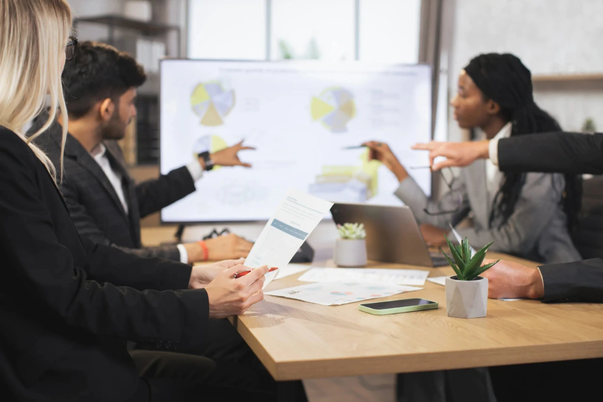 A group in business attire reviews a presentation with pie charts on a screen at a table with a laptop and paperwork.