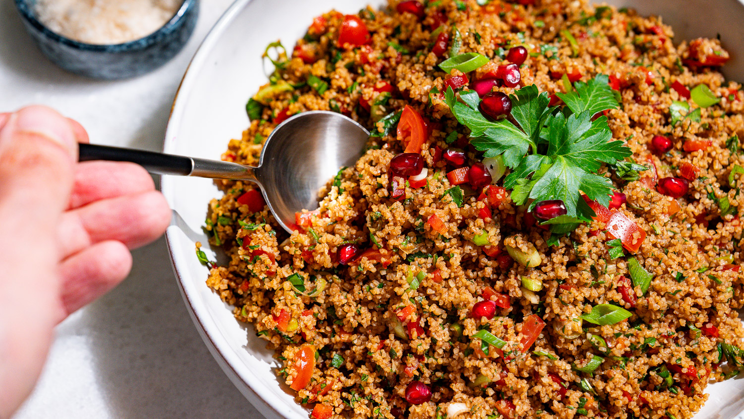 One hand is helping itself with a spoon from a bowl of kisir - Turkish bulgur salad.