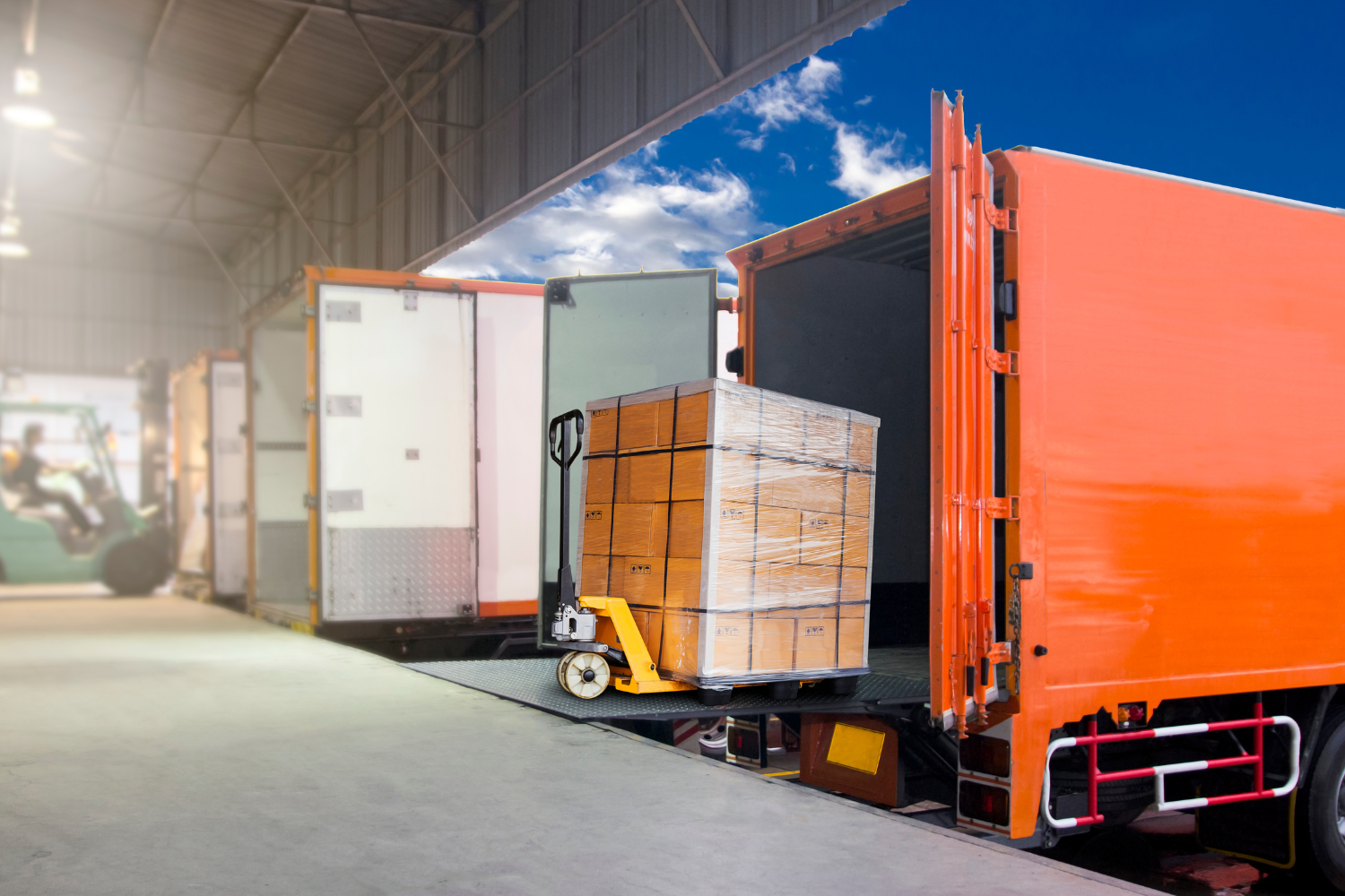 A pallet jack with a stack of boxes wrapped in plastic is positioned on a loading ramp next to an open orange truck trailer.