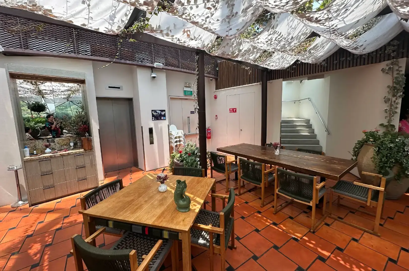 A quiet corner of a dining space with terracotta tiled flooring, displaying a long wooden communal table, an adjacent four-person dining set, an elevator entrance, and a staircase beneath draped white overhead canopy fabric.