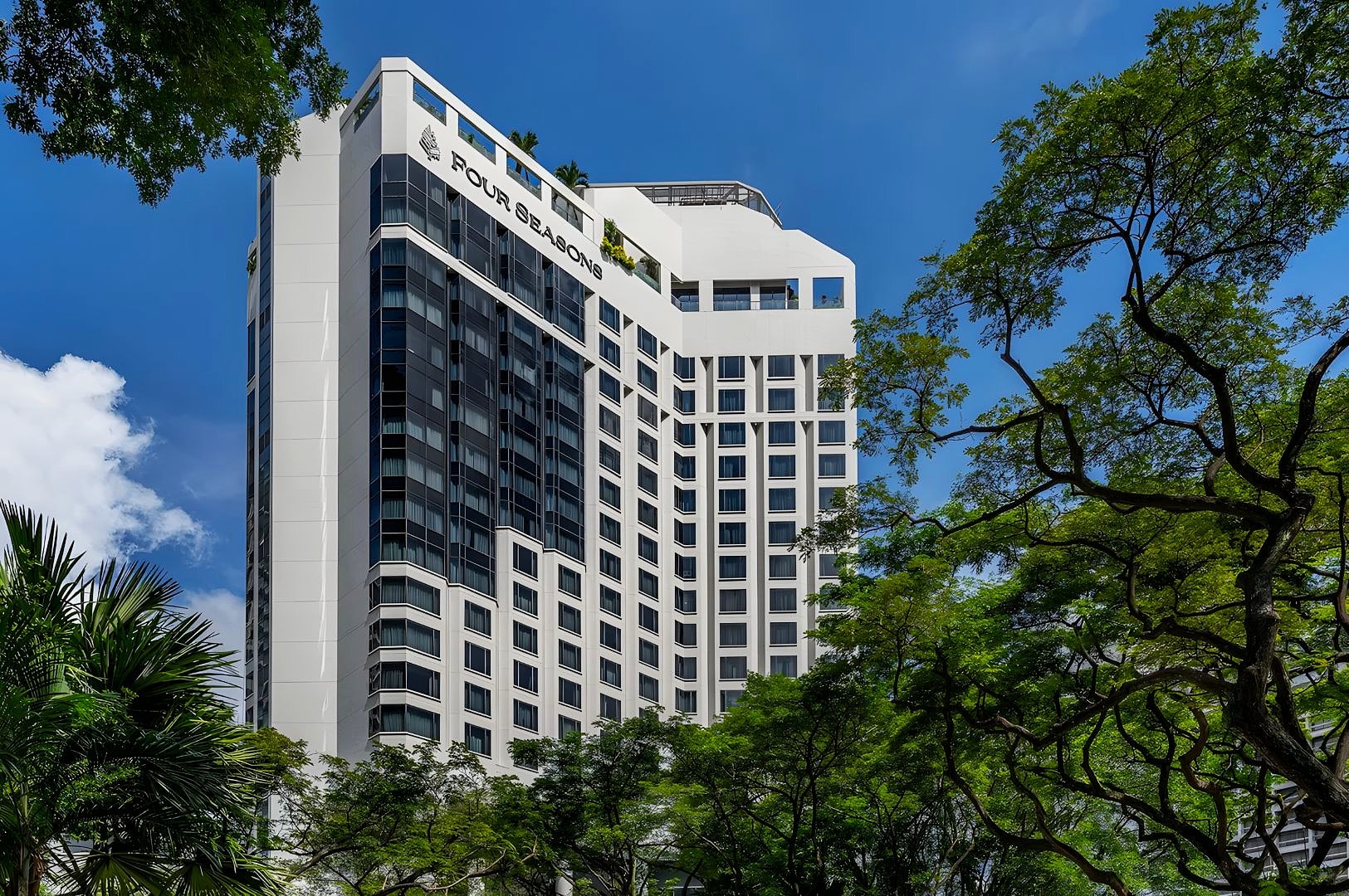 Tall, elegant hotel building with a modern design, framed by lush green trees under a clear blue sky, creating a serene and inviting atmosphere.