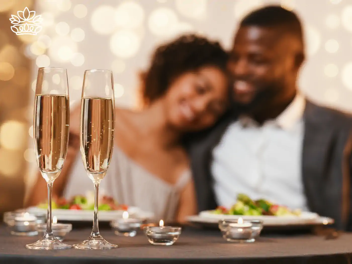 A couple enjoying a romantic dinner with champagne glasses in the foreground, illuminated by soft, twinkling lights, embodying a warm, celebratory Valentine's Day atmosphere. Fabulous Flowers and Gifts delivered with heart. Valentine's Day Flowers.