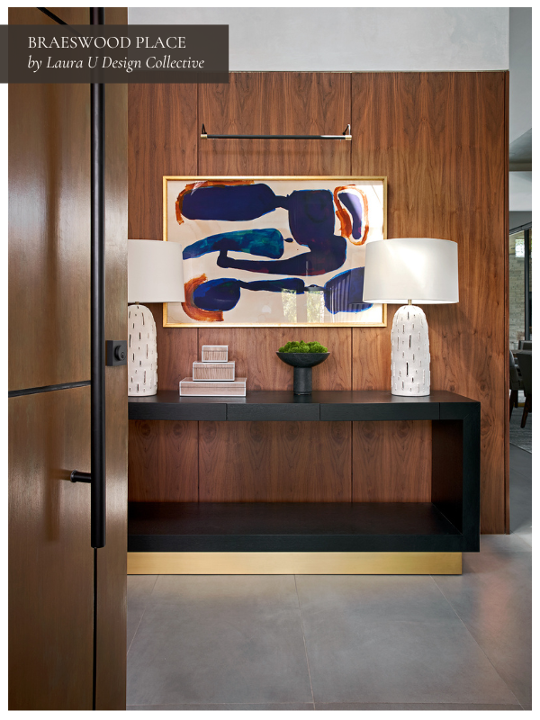A warm wood-paneled entryway with a bold abstract painting in blue and rust tones. A sleek black console with brass accents is styled with matching white ceramic lamps and stacked books.