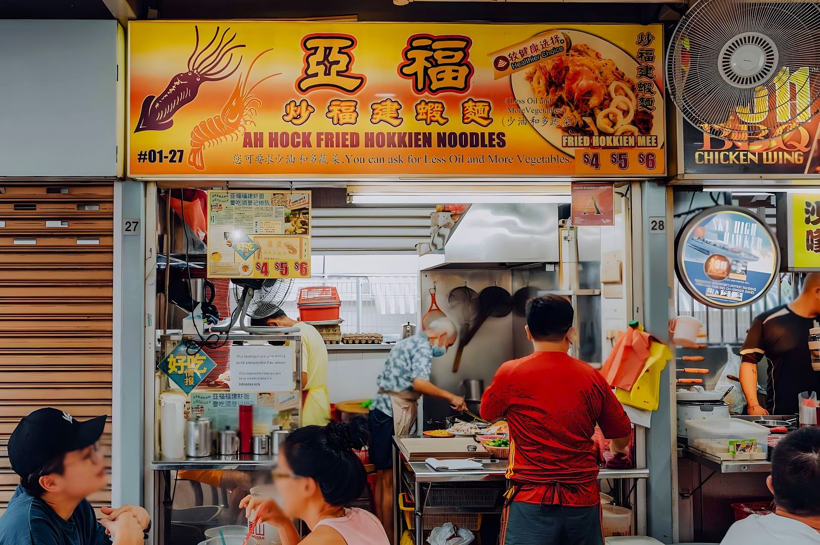A bustling hawker stall named "Ah Hock Fried Hokkien Noodles." A cook is preparing food in a small kitchen as customers wait. The atmosphere is lively.