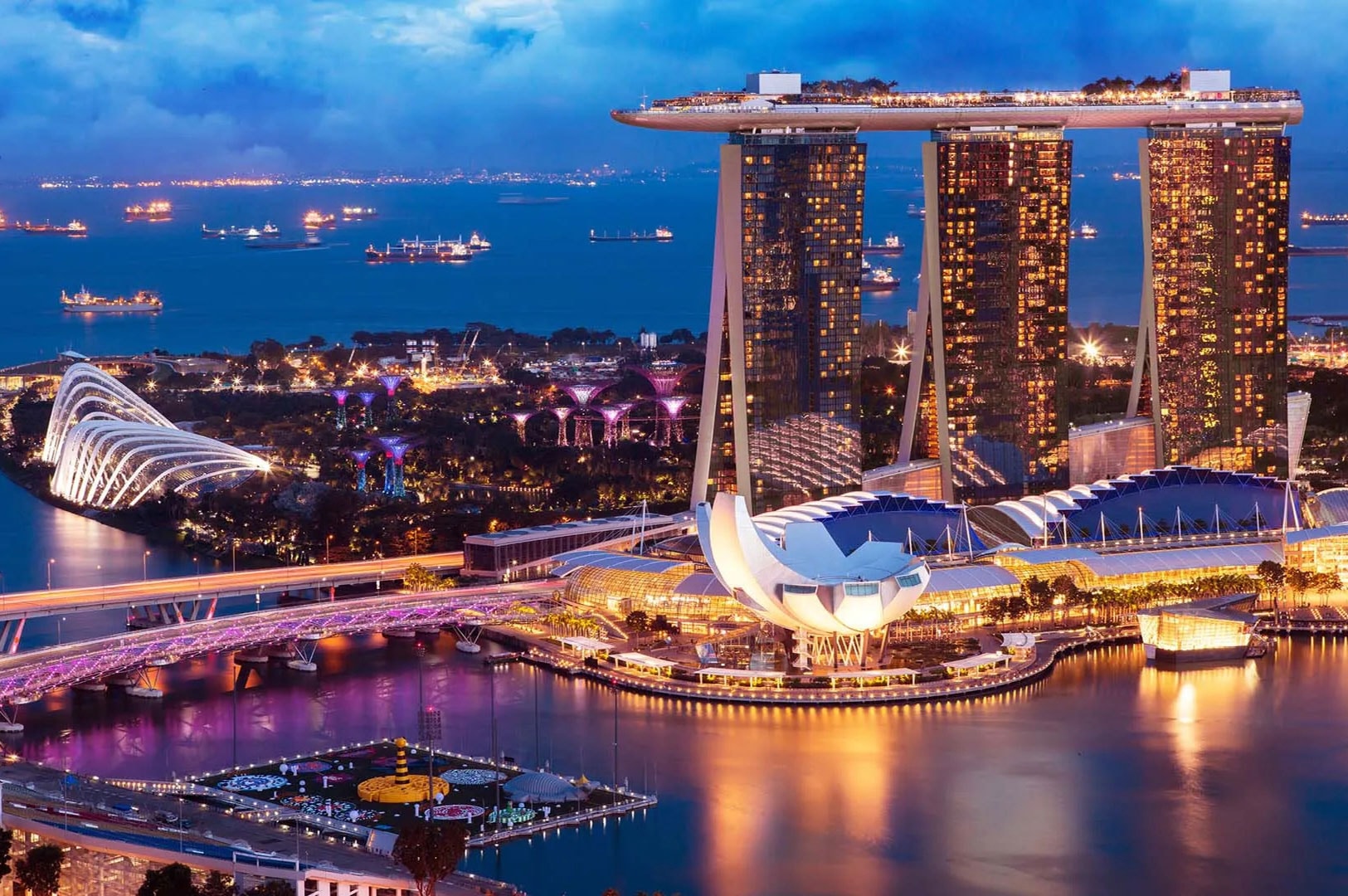 Nighttime cityscape of Marina Bay Sands in Singapore, featuring its three illuminated towers topped by the SkyPark, overlooking Gardens by the Bay, the Helix Bridge, and waterfront buildings reflecting colorful lights across Marina Bay, with ships visible in the harbor under a deep blue evening sky.