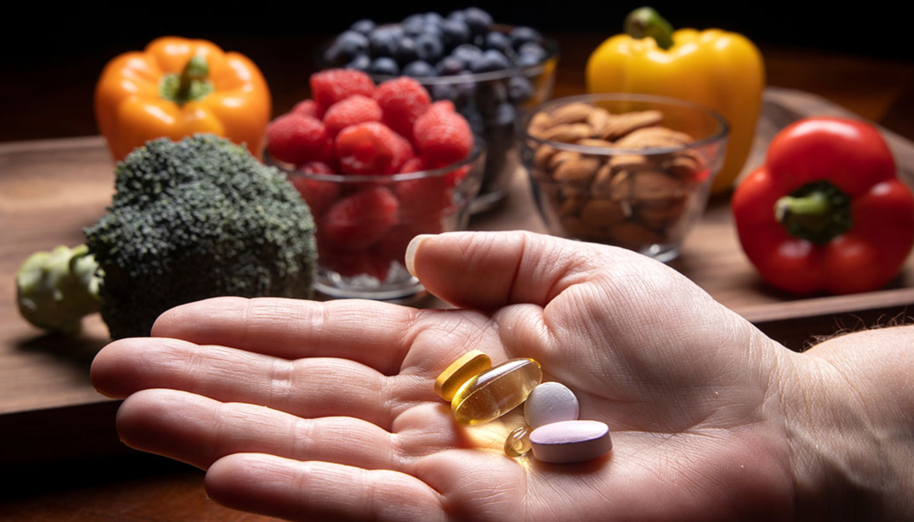 B-vitamin og tannhelse – Hva bør du vite? 12 Hånd med vitamintilskudd; frukt, grønnsaker og nøtter i bakgrunnen.