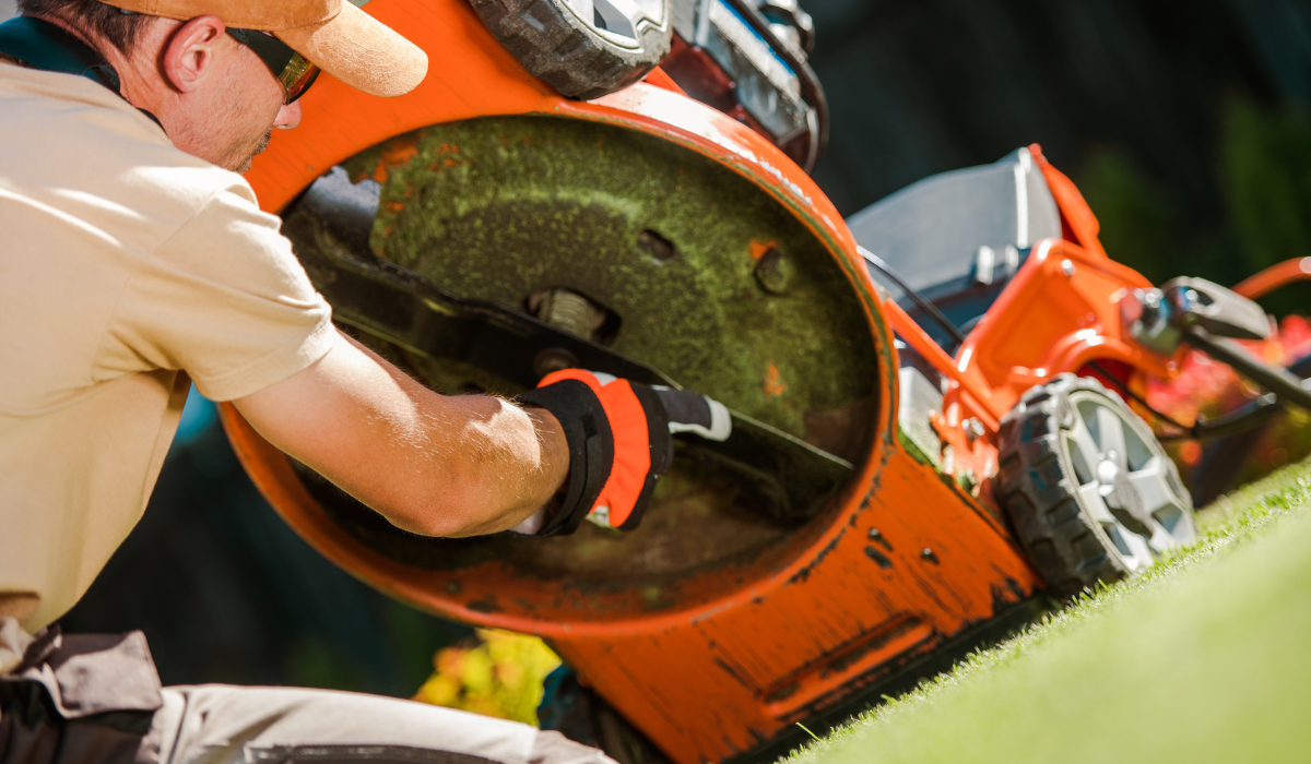A figure using a brush to clean dried grass from the underside of a rotary lawn mower