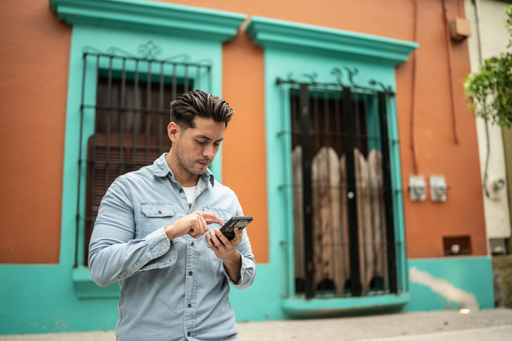Young man in a blue shirt sending a text in front of an orange and teal building.