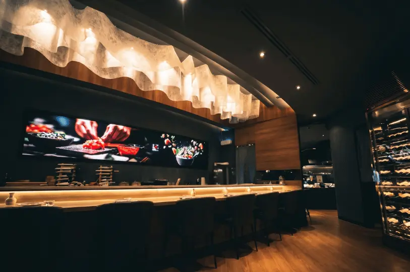 A modern, upscale izakaya setting featuring a long, illuminated wooden counter lined with dark chairs. A wavy, decorative light fixture hangs from the ceiling, and a large digital screen displays vibrant food preparation imagery on the back wall.