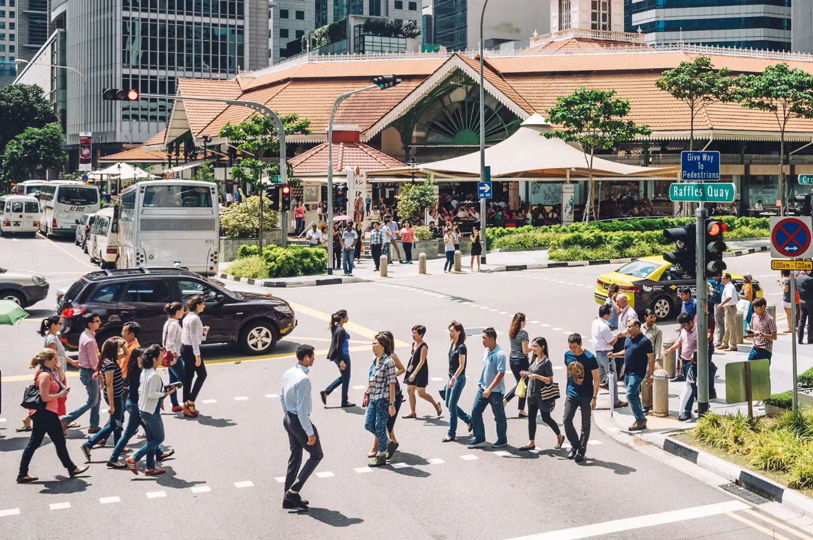 People crossing a busy street in a city with cars and buses, and buildings in the background.