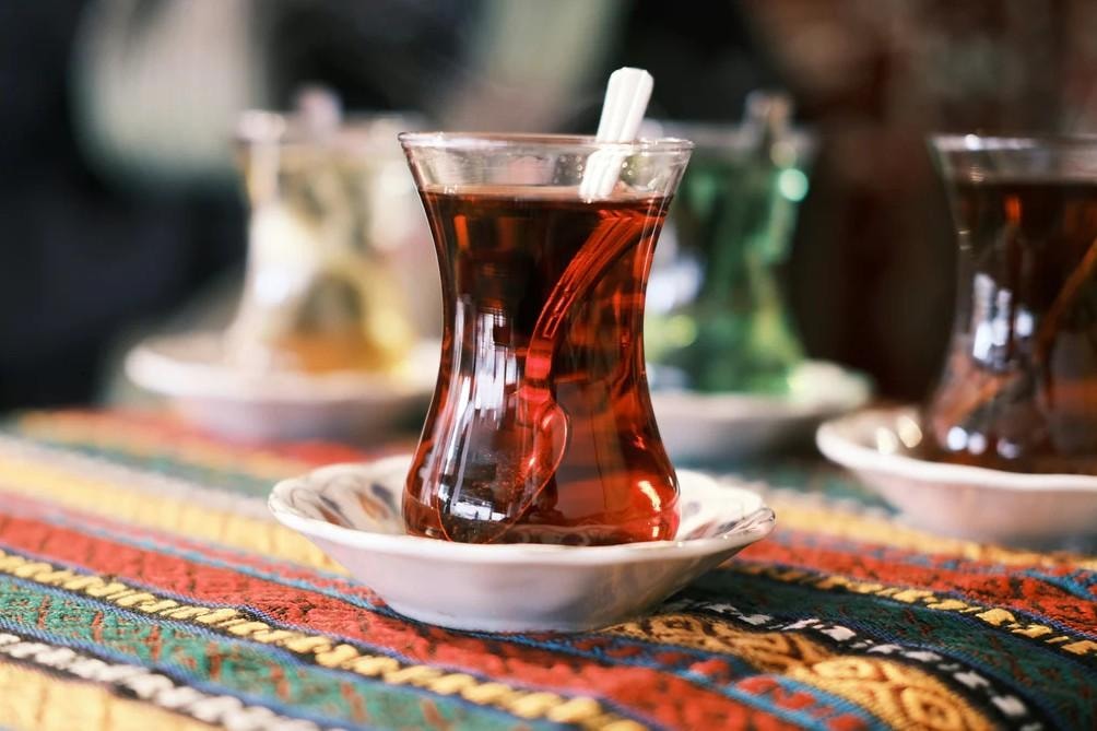 A traditional Turkish tea glass filled with dark amber tea sits on a white saucer atop a vibrant, patterned tablecloth. A small spoon is tucked inside the curved glass, while other tea servings remain softly blurred in the background.