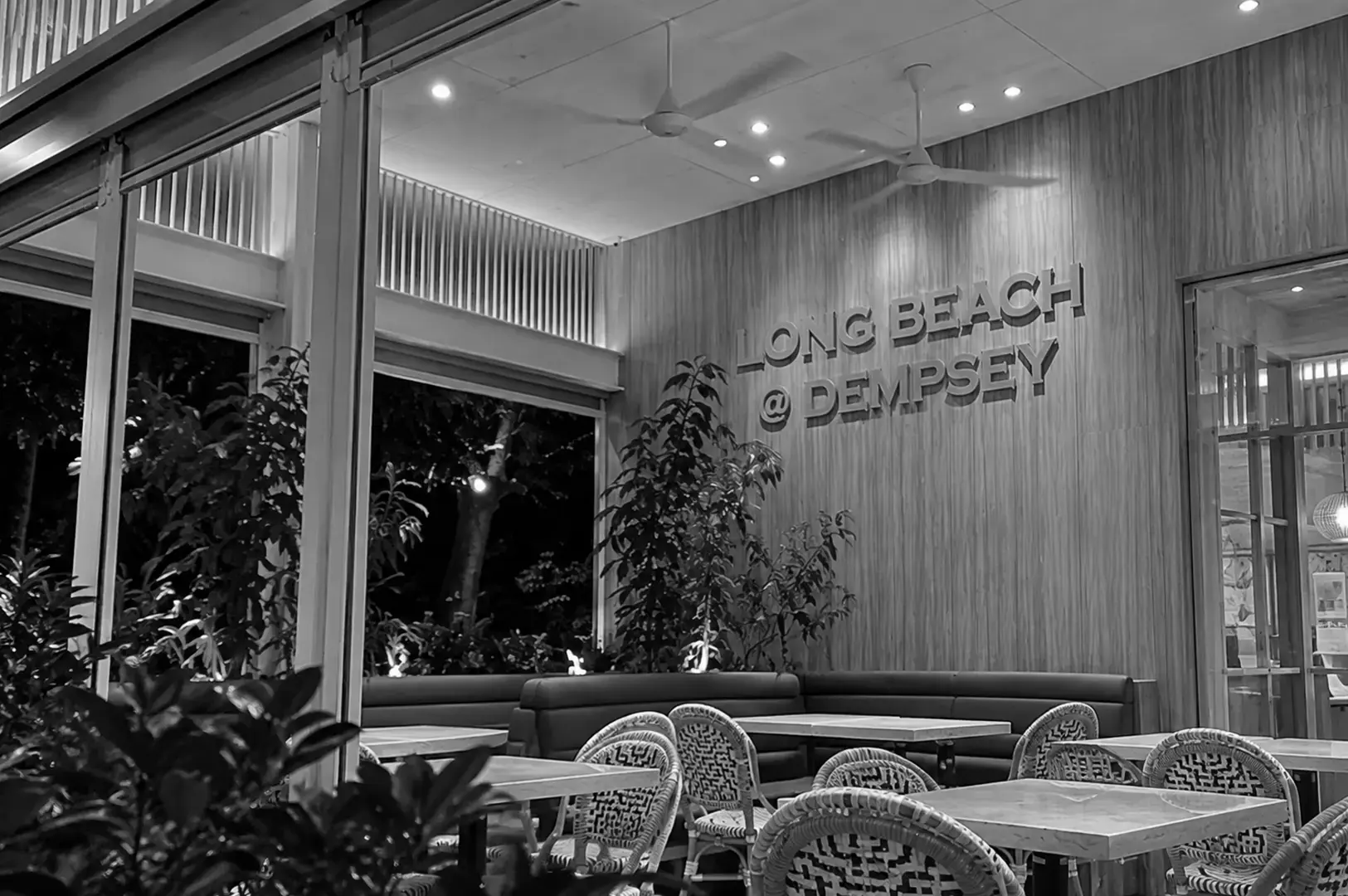Interior seating area of Long Beach at Dempsey, featuring wooden wall panels, ceiling fans, large open windows, woven chairs, and tables surrounded by greenery.
