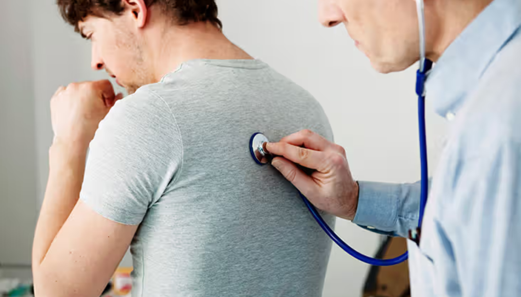 Lege lytter til en manns pust med stetoskop mens pasienten hoster og sitter med ryggen vendt mot legen i et undersøkelsesrom.