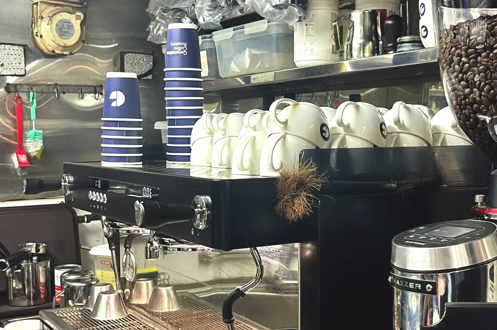 A close-up of a professional commercial espresso machine, stacked blue takeaway cups, white ceramic mugs, and a hopper filled with dark coffee beans.