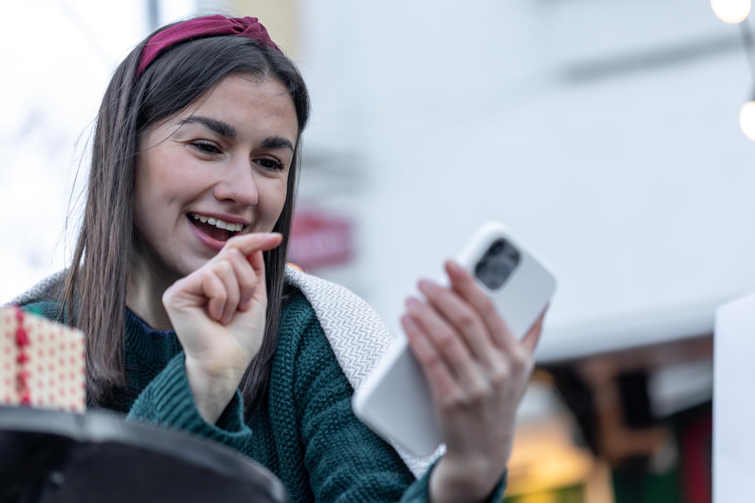 A young woman with a maroon headband and green sweater smiles brightly while video chatting or texting on her smartphone in an outdoor café. Her expressive gesture underscores the human connection enabled by mobile messaging solutions.