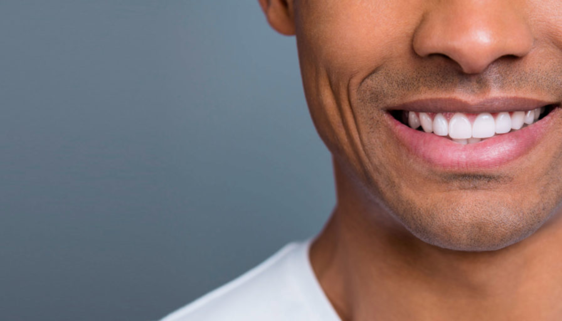 Nærbilde av smilende mann med hvite tenner og glatt hud foran nøytral, grå bakgrunn.