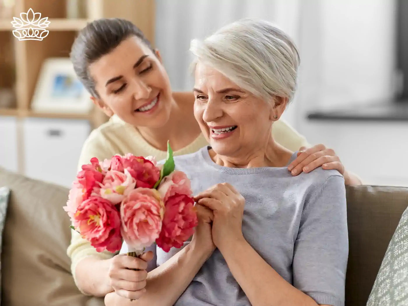 A younger woman presents a bouquet of lush pink peonies to an elderly woman, sharing a moment of joy and affection in a cosy living room setting. Fabulous Flowers and Gifts delivered with heart. Valentine's Day Flowers.