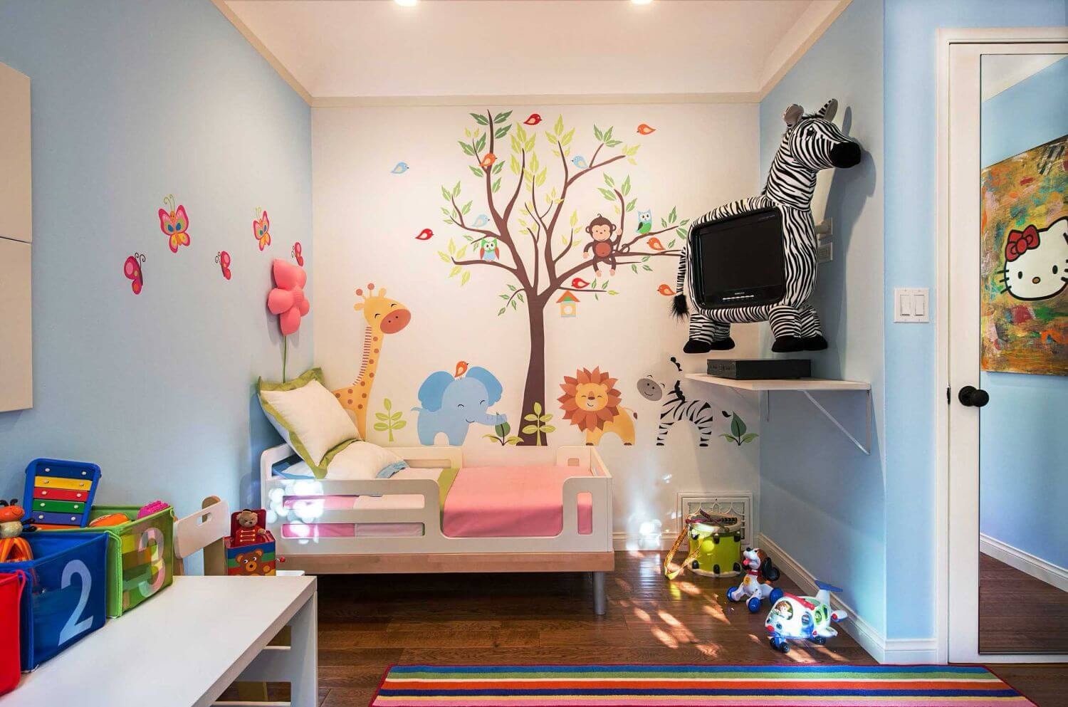A vibrant toddler room features pastel blue walls adorned with playful animal-themed decorations, creating an inviting and imaginative space for kids. The room is designed with storage solutions, television, and colourful bedding, perfect for a little guy/girl's bedroom decor.
