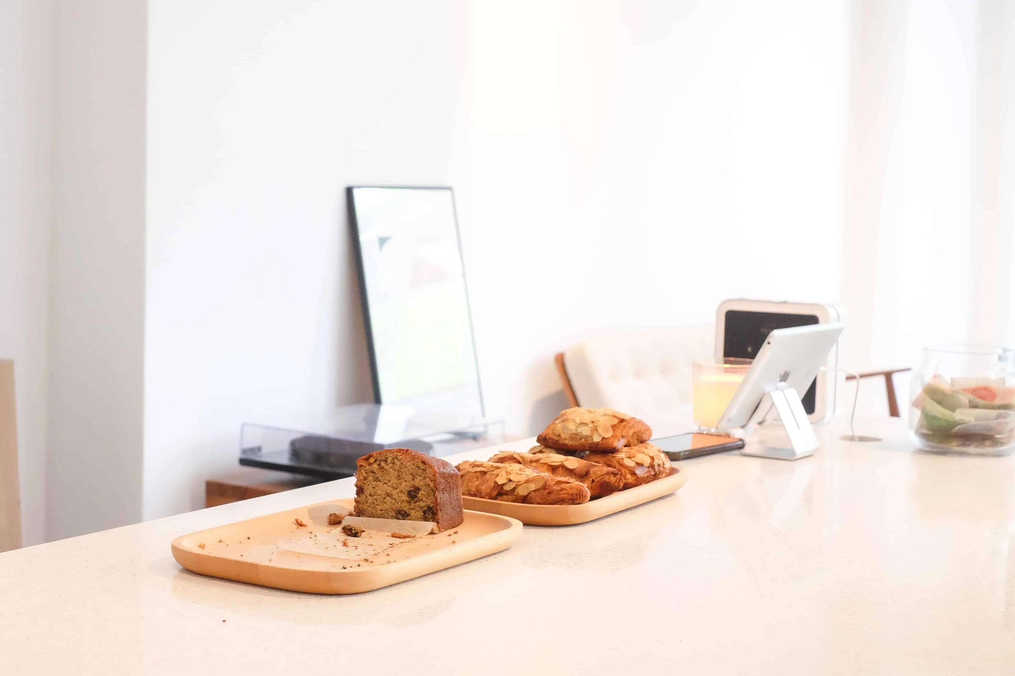 A slice of banana bread and several almond croissants sit on wooden trays atop a bright, minimalist white counter. The background features a clean, airy aesthetic with a tablet on a stand and a glass jar of snacks in soft, natural light.