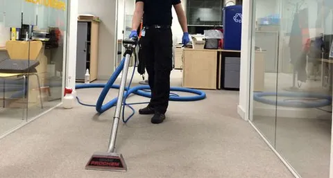 A professional cleaner using a Prochem carpet steam cleaning machine on office carpeting, with long blue hoses extending through a glass-partitioned corridor.