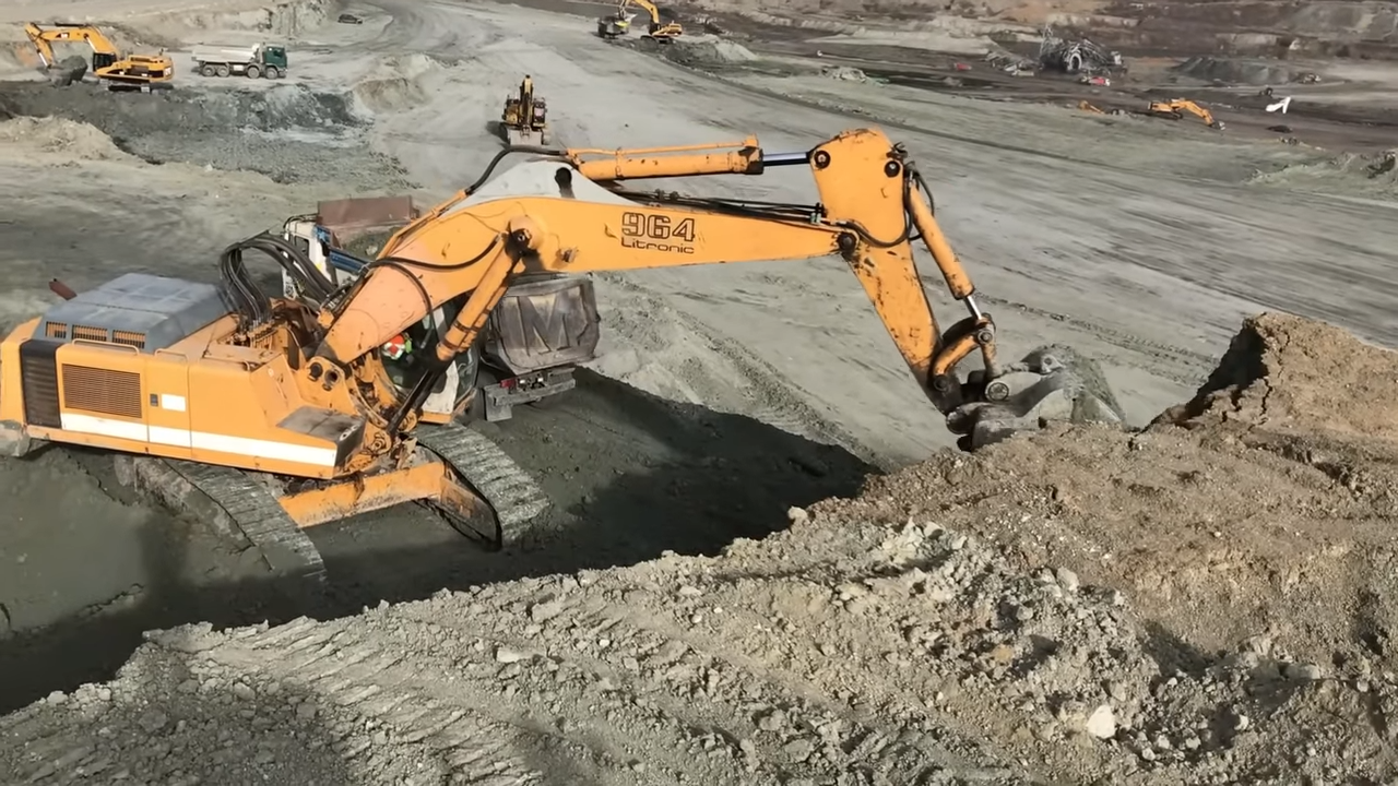 A large orange excavator with 964 Litronic written on its arm is digging and moving dirt at a construction site, with several other heavy machinery and trucks in the background