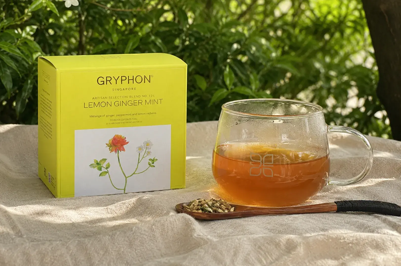 A bright yellow box of Gryphon "Lemon Ginger Mint" tea sits on a textured, off-white linen tablecloth with a soft-focus background of sun-dappled green leaves. Next to the box, a clear, wide-bottomed glass mug is filled with a warm, golden-orange herbal tea, casting a soft glow onto the fabric. In the foreground, a slender wooden spoon with a black-wrapped handle lies flat, holding a small portion of loose, dried herbal tea blend consisting of cut ginger bits and mint leaves.
