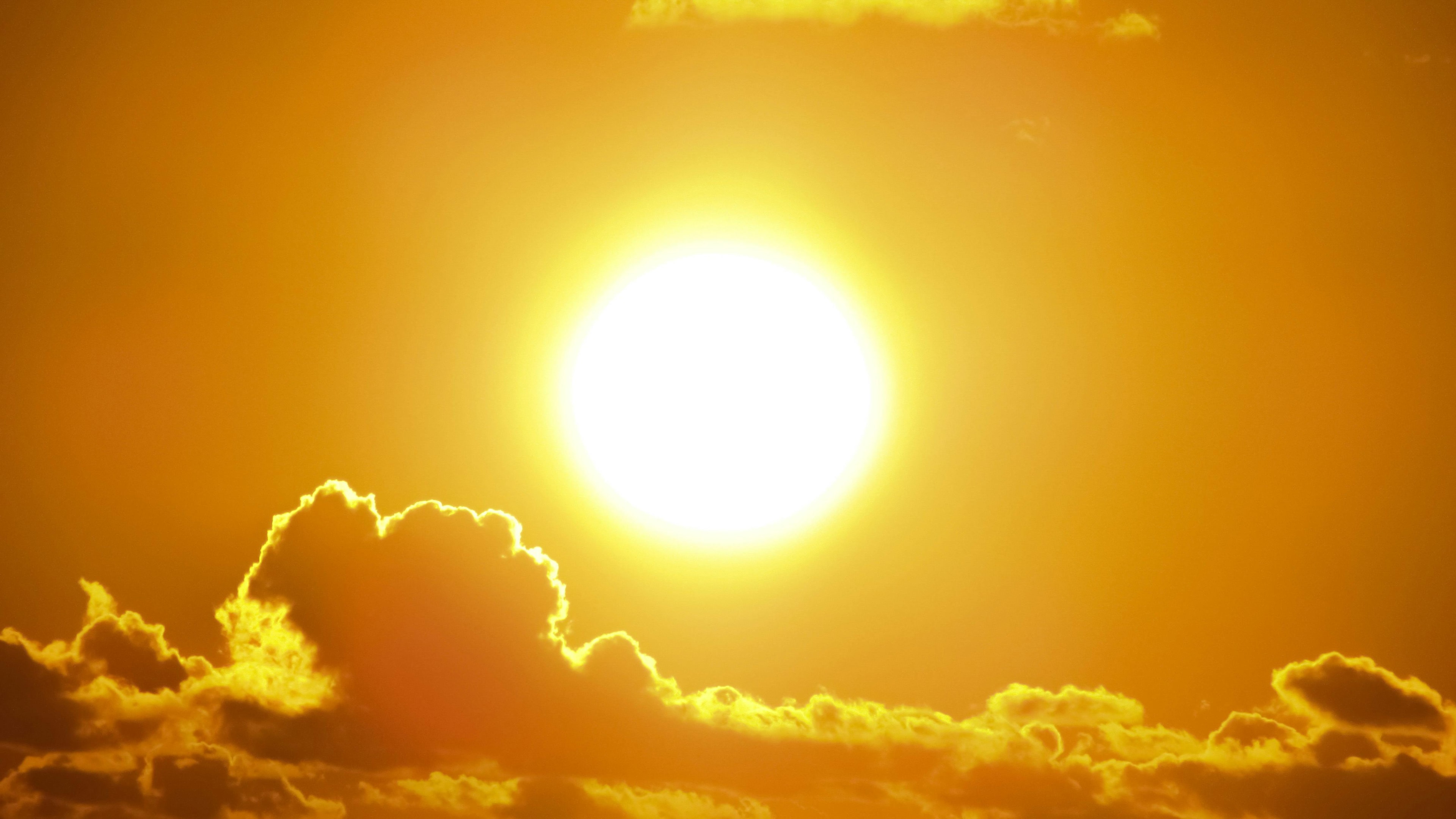 A bright, glowing sun in a clear yellow sky, partially obscured by a few clouds, representing intense sunlight and heat.