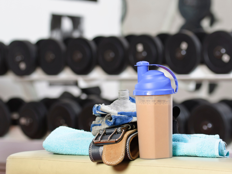 An image showing protein powder and a shaker bottle on a gym bench.