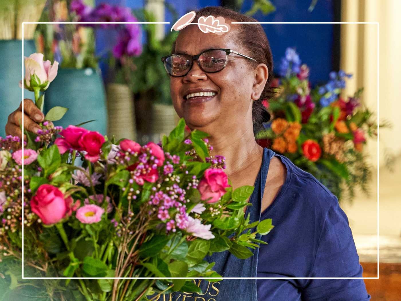 A woman in a dark blue shirt and glasses smiling joyfully as she holds a vibrant bouquet with pink roses and assorted flowers, in a lively floral shop. Fabulous Flowers and Gifts.