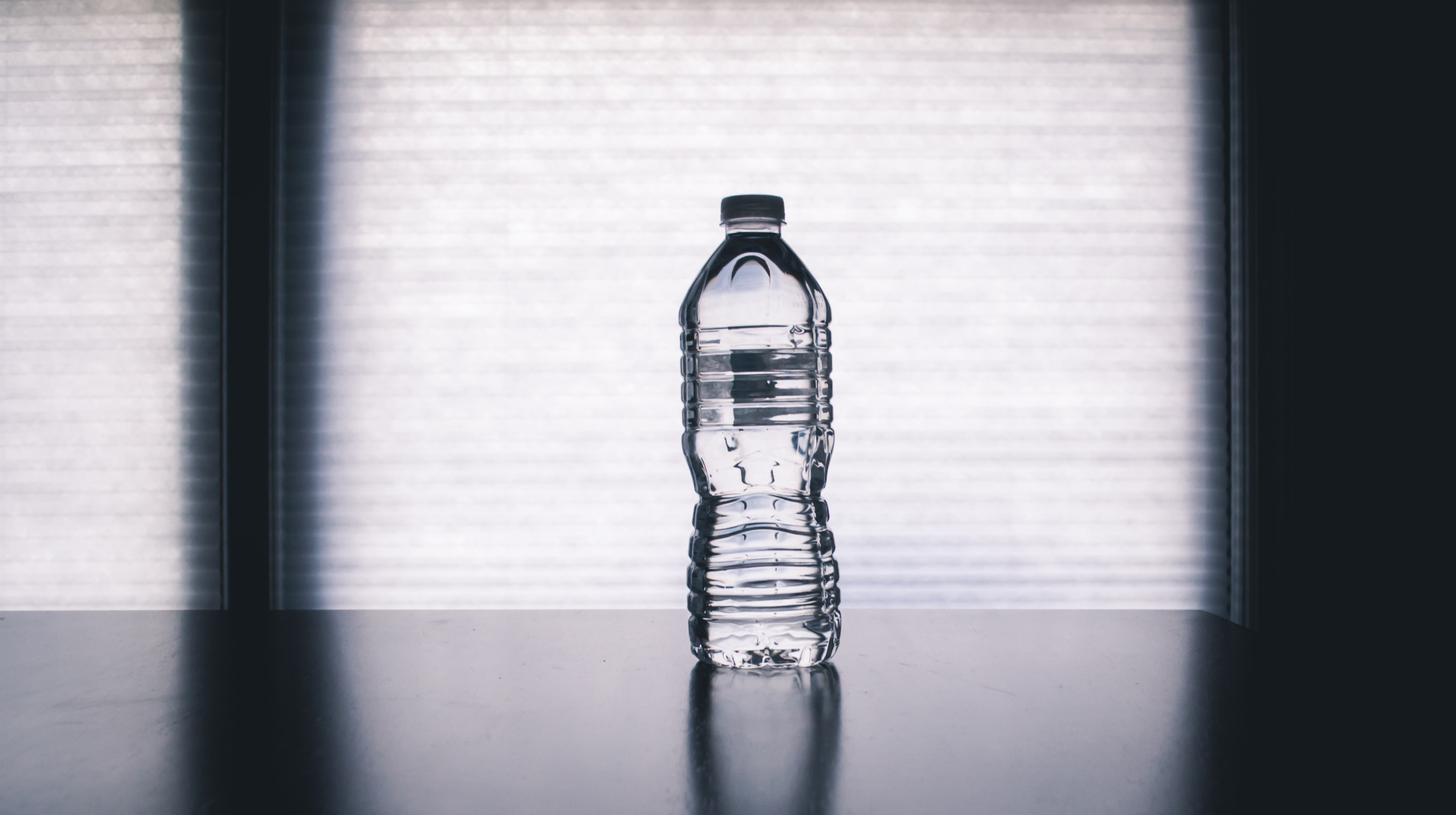 Une bouteille d'eau en plastique pleine et fermée est posée sur une table dans une salle un peu obscure. On distingue une large fenêtre derrière la bouteille.