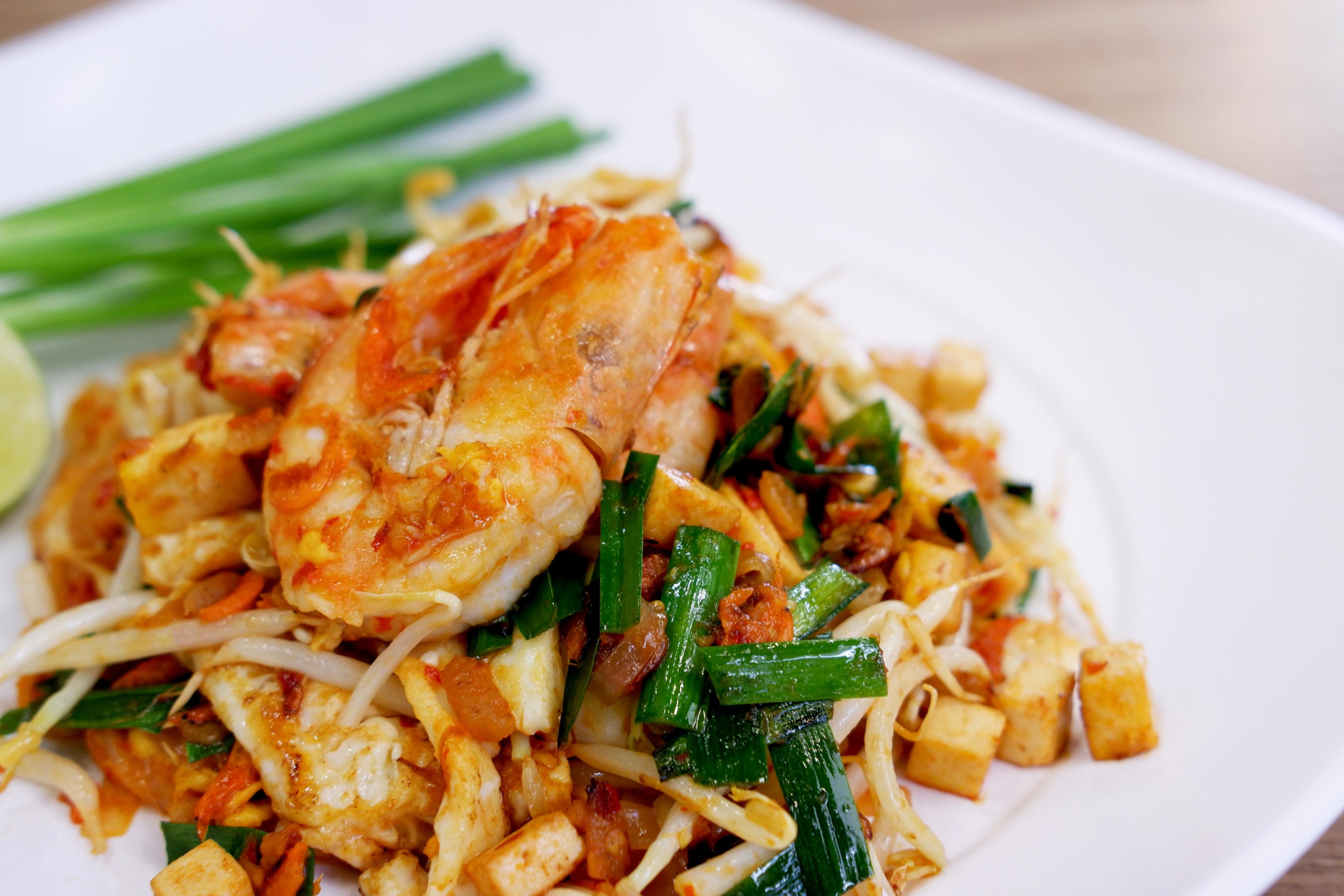 This image features a vibrant plate of Pad Thai, showcasing stir-fried rice noodles tossed with succulent shrimp, golden cubes of tofu, and fresh bean sprouts. Garnished with bright green garlic chives and a wedge of lime on the side, the dish is presented on a clean white plate for a fresh, appetizing look.