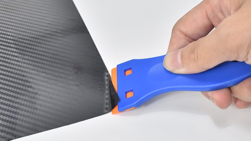 blue plastic scraper with orange tip removing the carbon fiber vinyl wrap from the surface