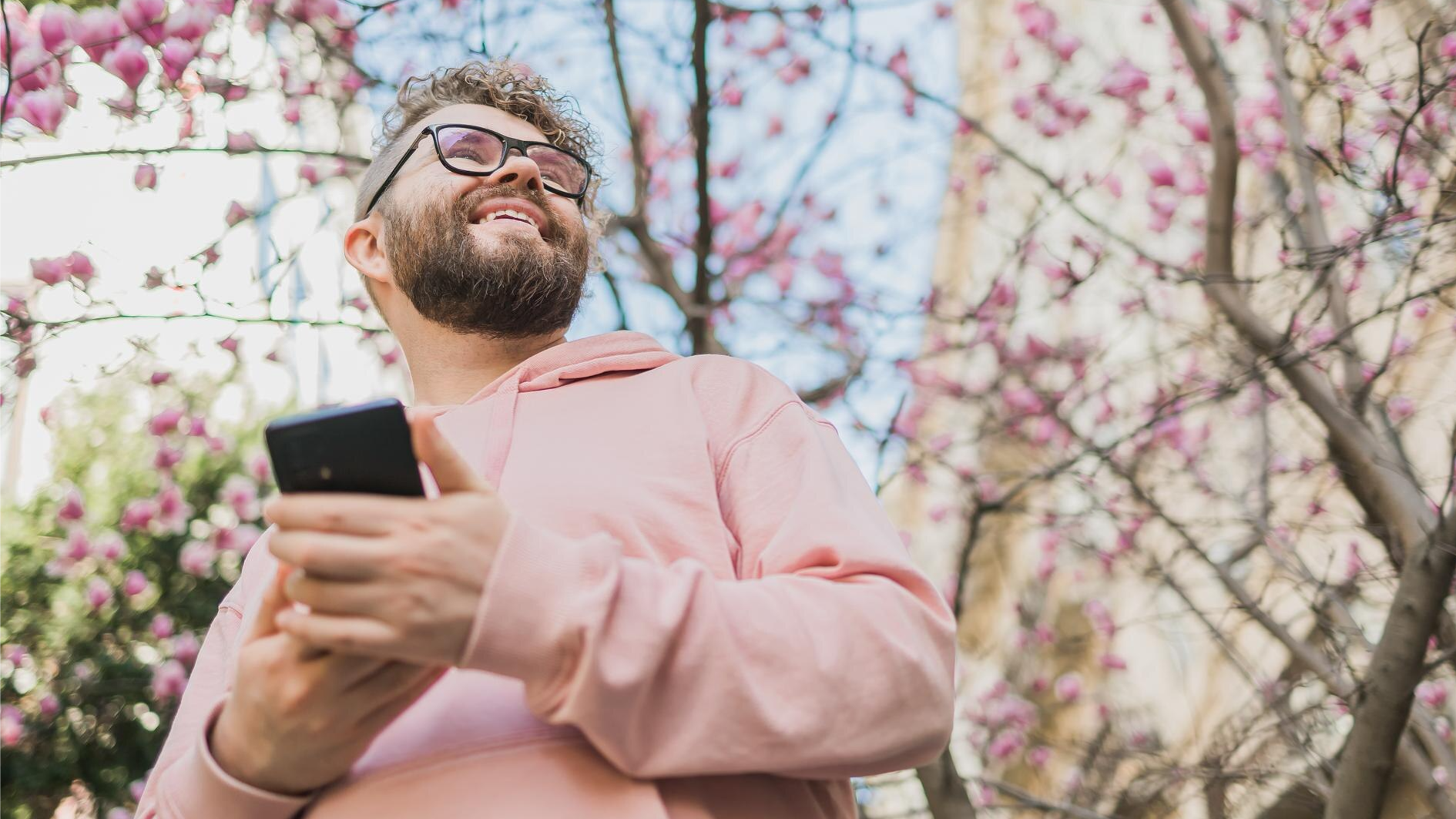 A smiling man in a pink hoodie holds a smartphone while standing outdoors near blooming pink magnolia trees. His joyful expression and mobile device highlight the use of business SMS and mobile communication in a relaxed, modern work environment.