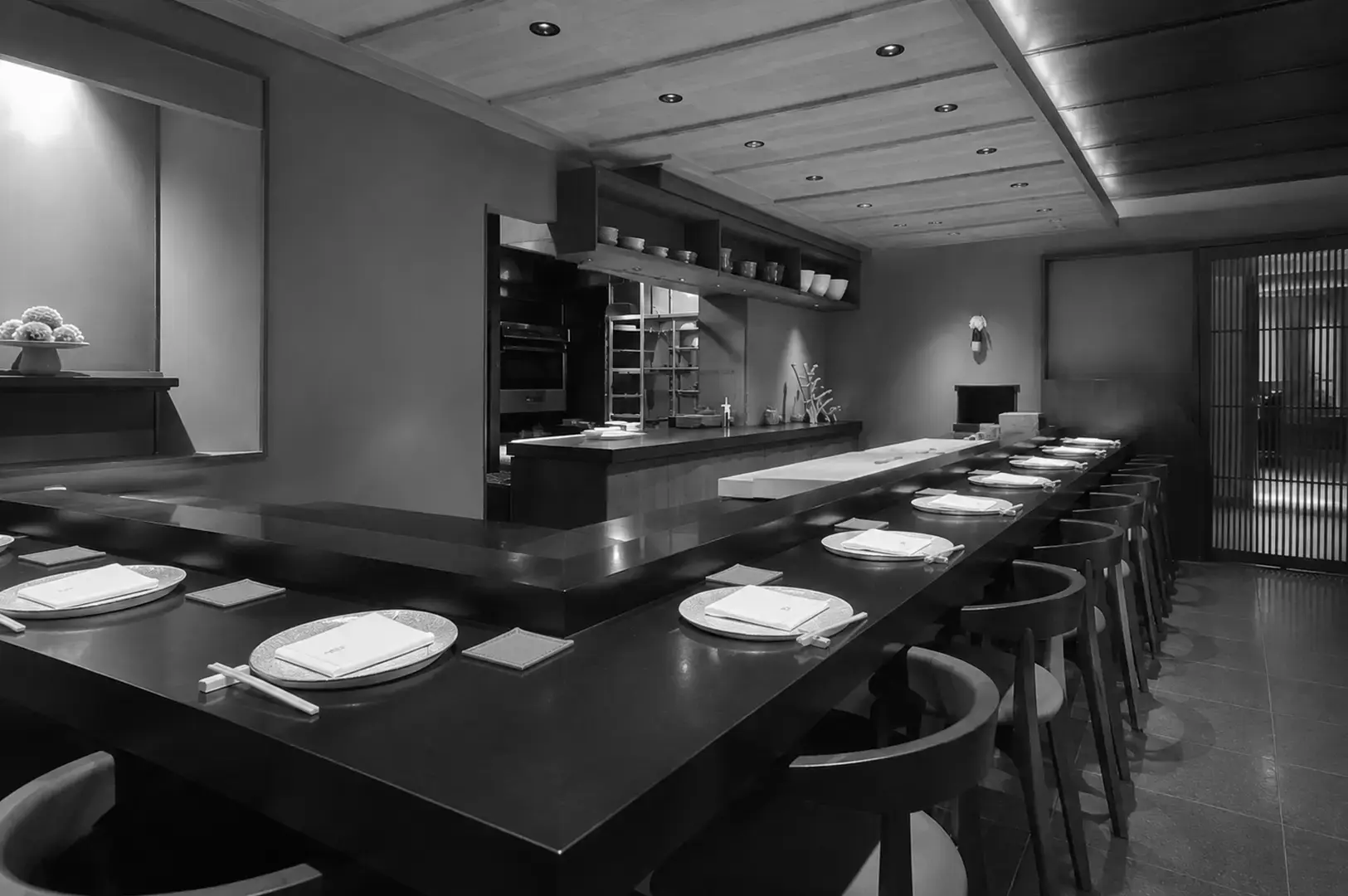 Modern Japanese sushi bar with long dark counters, neatly arranged table settings, open kitchen workspace, and warm recessed lighting.