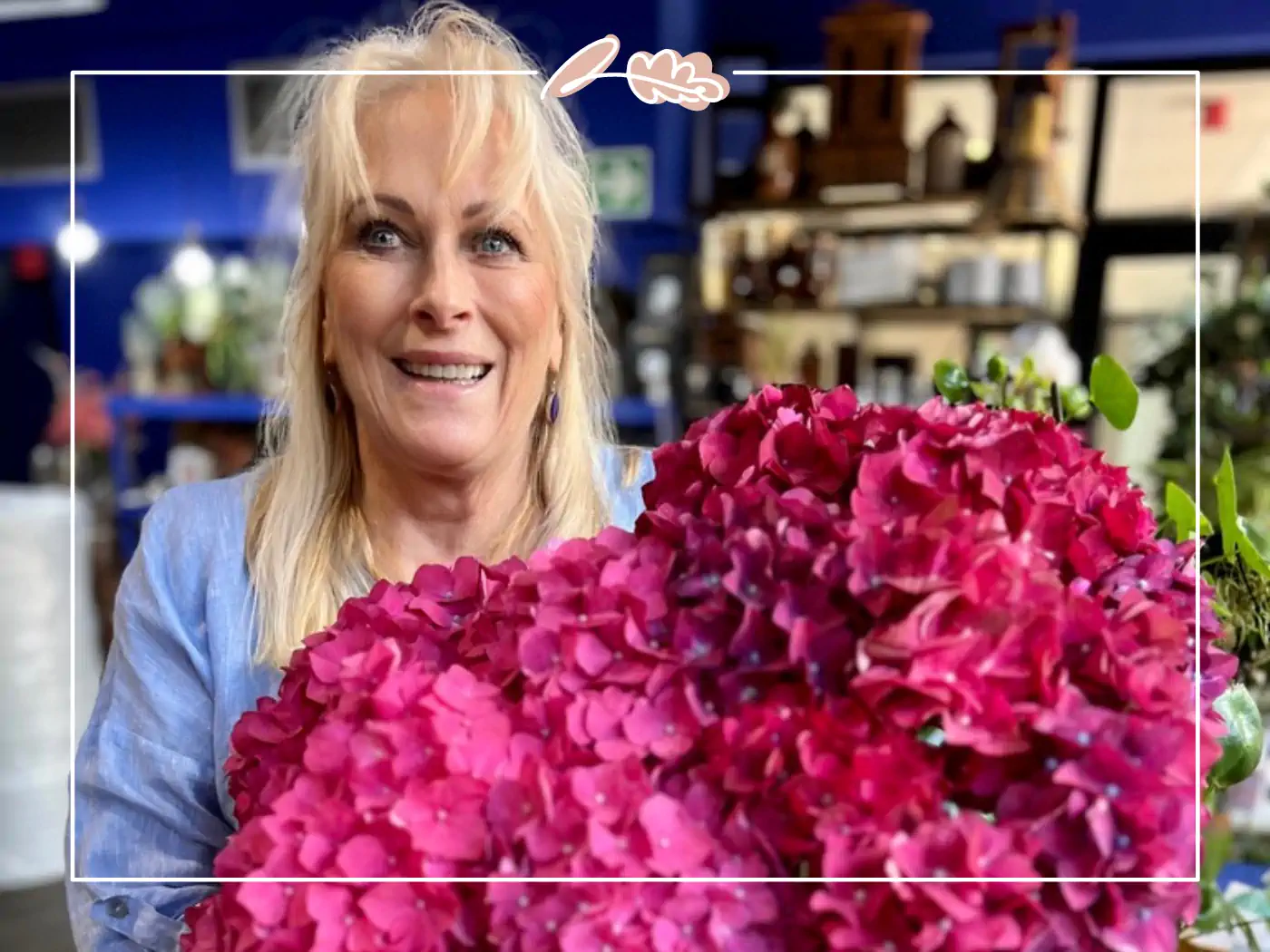 A woman with blonde hair in a blue shirt, holding a pink bouquet of hydrangeas, smiling.