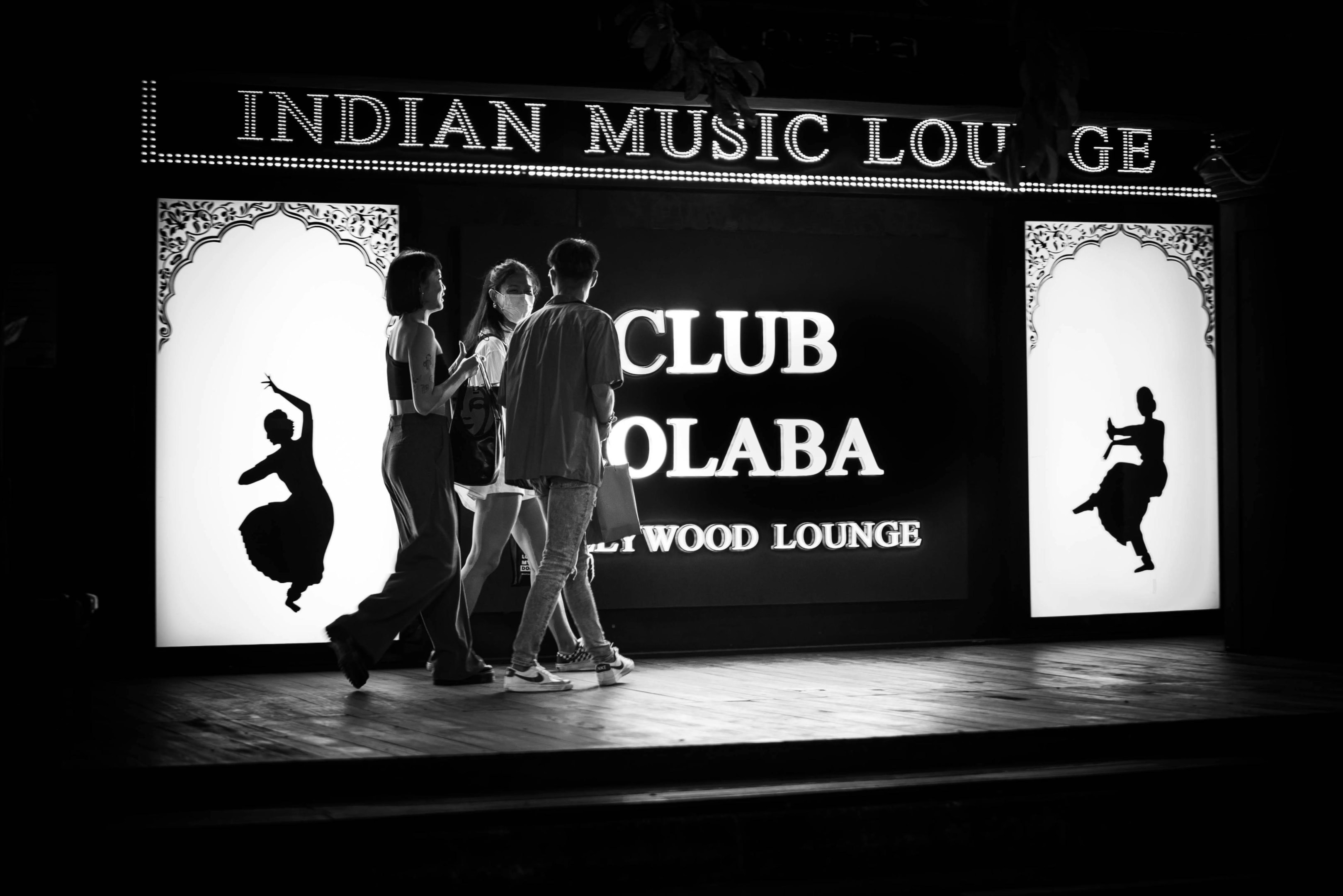 Three pedestrians stroll past a brightly illuminated sign for "Club Colaba," which also advertises itself as an "Indian Music Lounge" and "Bollywood Lounge". The black-and-white image captures the glowing display, featuring silhouette figures of traditional dancers, as the group walks across the wooden platform in front of the venue.