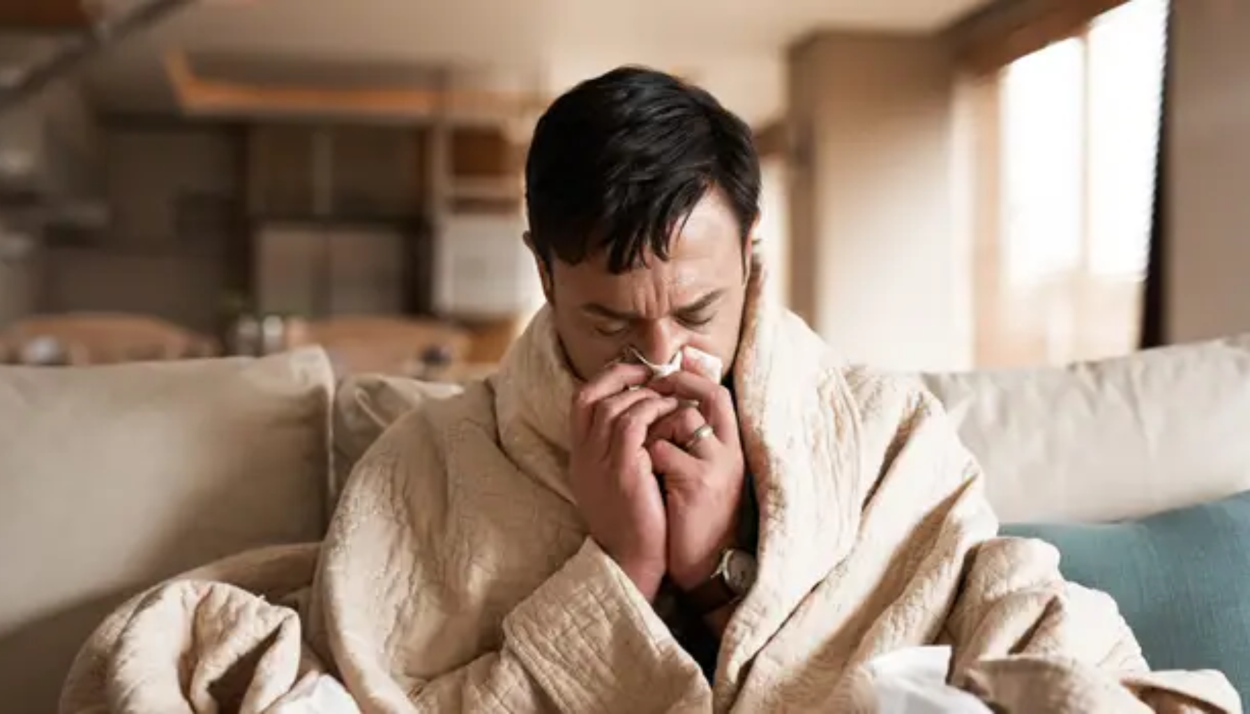 Mann pakket inn i varm genser sitter på sofaen og snyter seg i et papir.